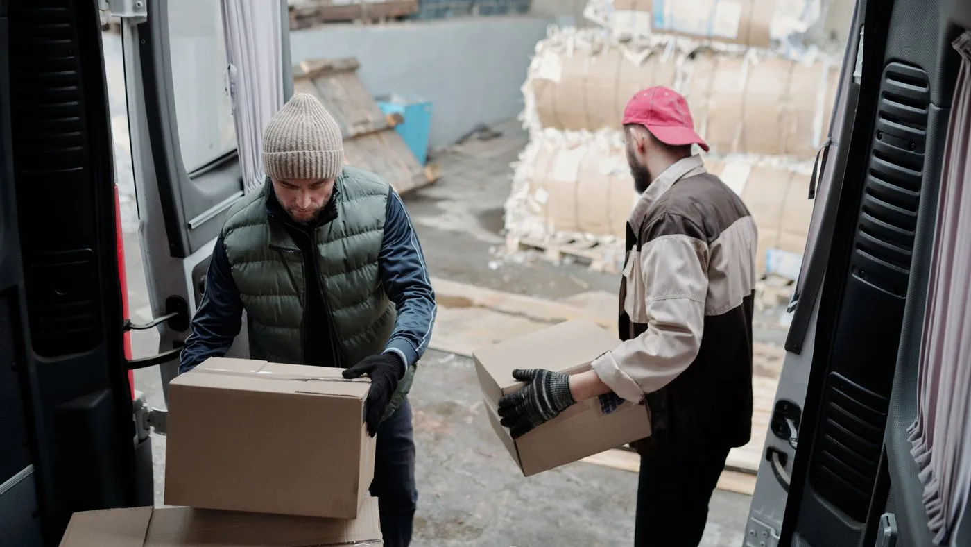 Two workers in gloves and body warmers passing cardboard boxes into the open rear doors of a van in an outdoor yard