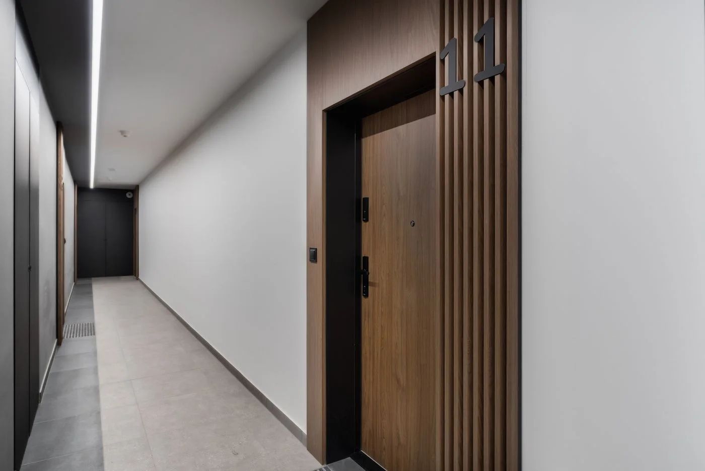 Modern apartment corridor with pale tiled flooring, white walls and a wood-panelled front door marked with the number 11