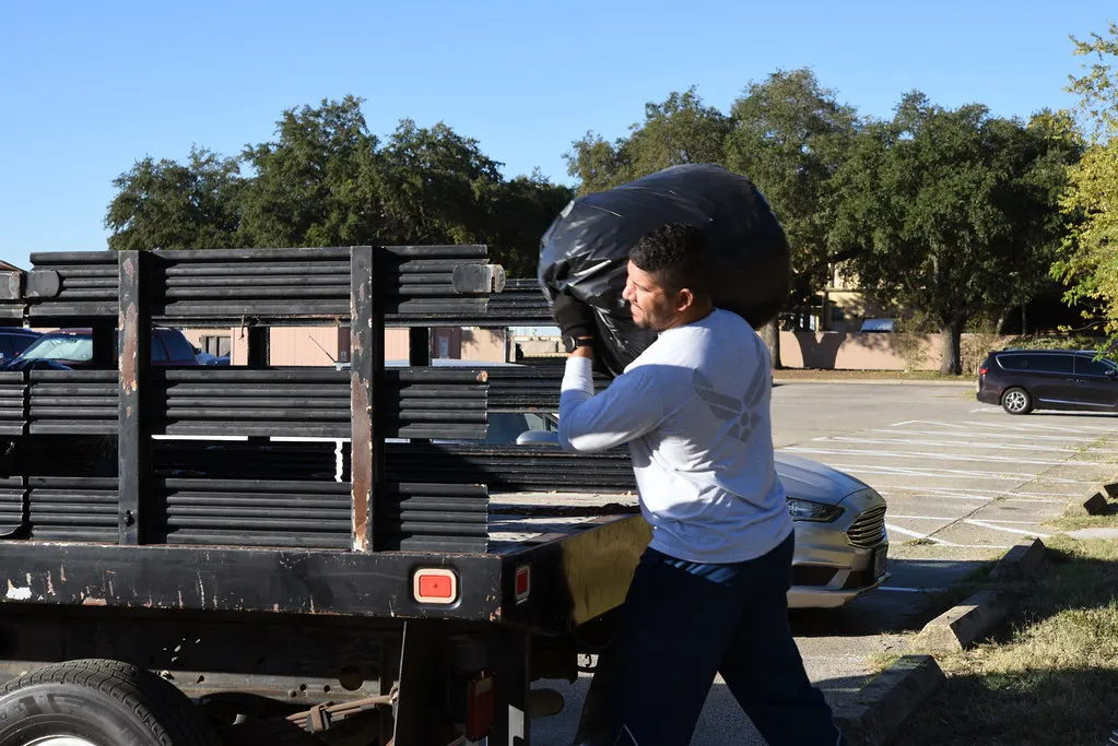 Worker in a long-sleeved top and gloves lifting a full black waste sack onto the bed of a black flatbed truck in a car park