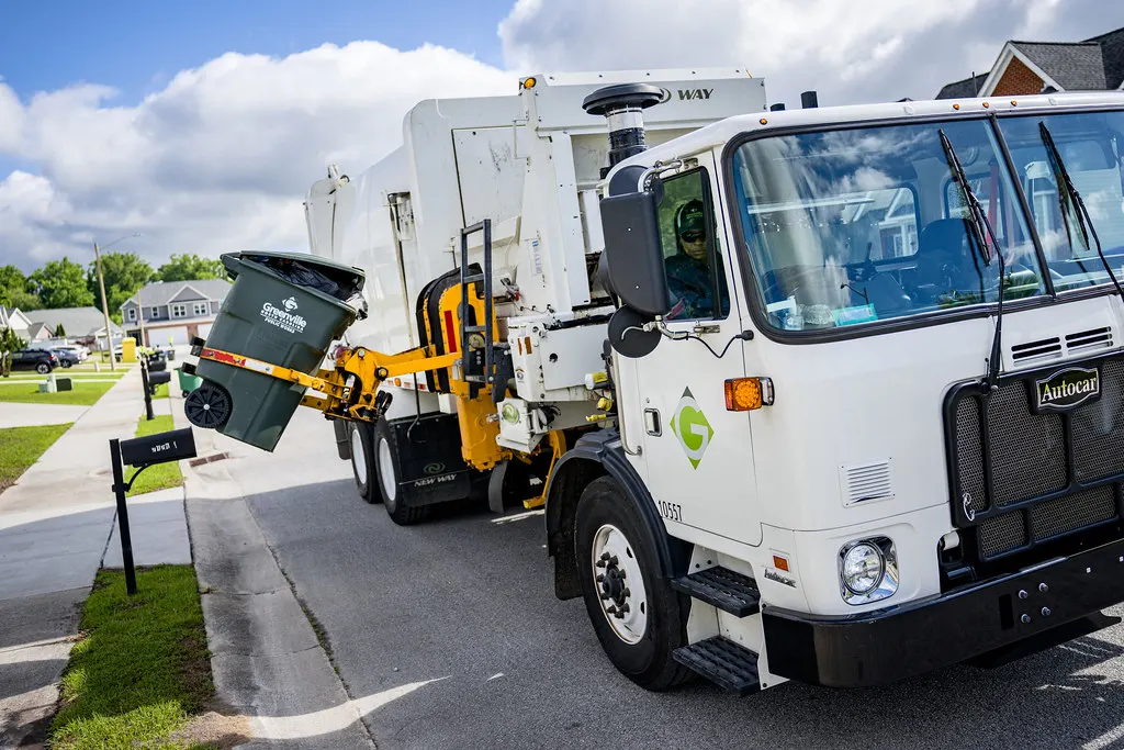 White waste collection truck with a mechanical side arm lifting a green wheeled bin at the kerbside of a suburban residential street