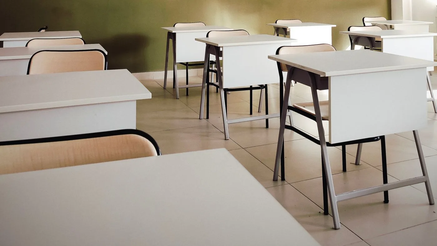 Empty classroom with rows of white study desks and pale wood chairs on tiled flooring against an olive green wall