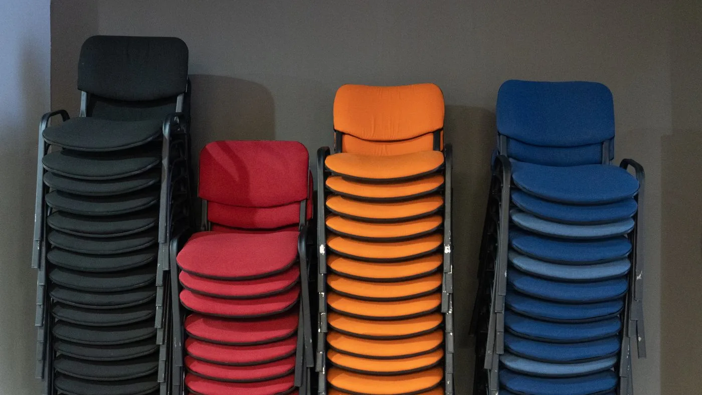Four tall stacks of upholstered stacking chairs sorted by colour, black, red, orange and blue, against a grey wall
