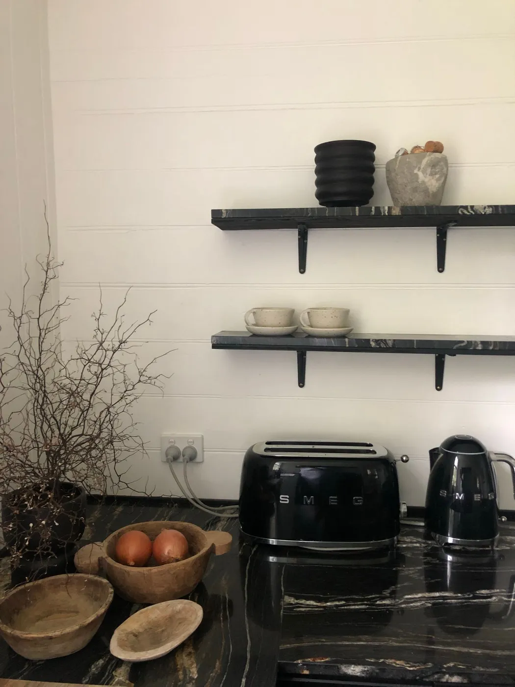 Black SMEG toaster and kettle on a dark marble kitchen counter beneath open shelving holding a black pot and two ceramic cups