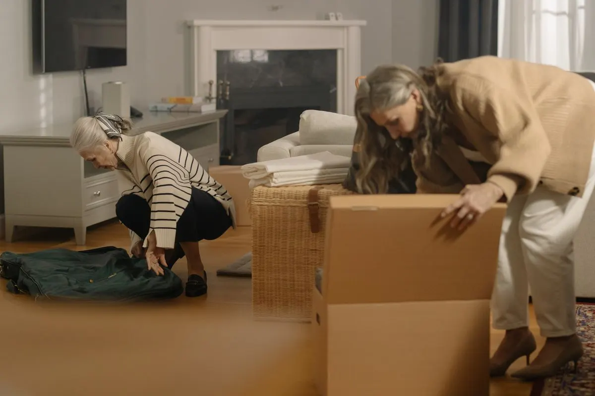 Two senior women packing cardboard boxes together in a living room with a fireplace