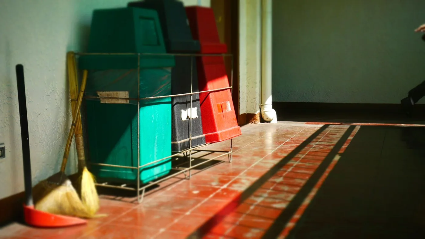 Green, black and red waste containers standing in a metal frame against a wall in a tiled building corridor, with two brooms leaning beside them