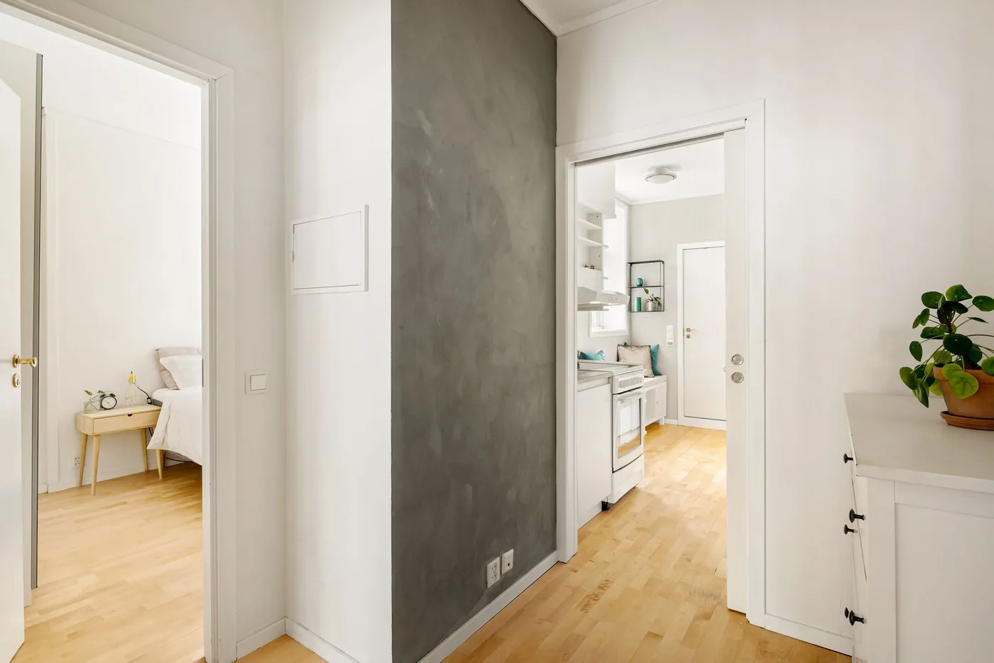 Bright apartment hallway with white walls and pale wood flooring, opening into a bedroom on one side and a compact white kitchen on the other