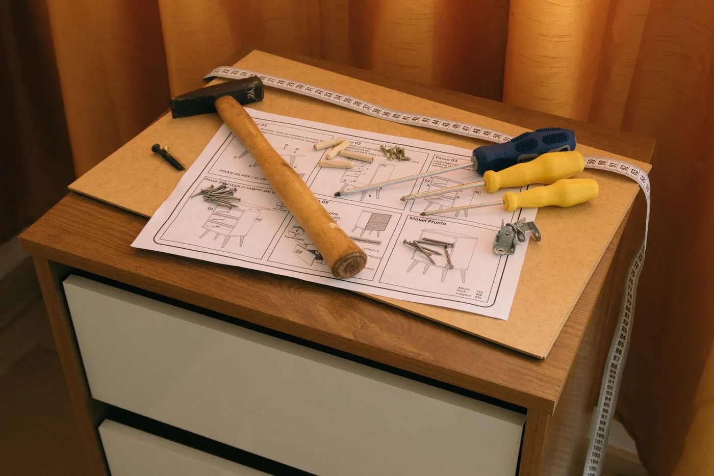 Part-assembled flat-pack cabinet with a printed instruction sheet, hammer, two screwdrivers, loose screws and a tape measure resting on its top panel