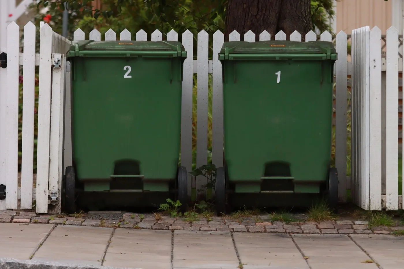 Two green wheelie bins numbered one and two standing beside a white picket fence outside a house