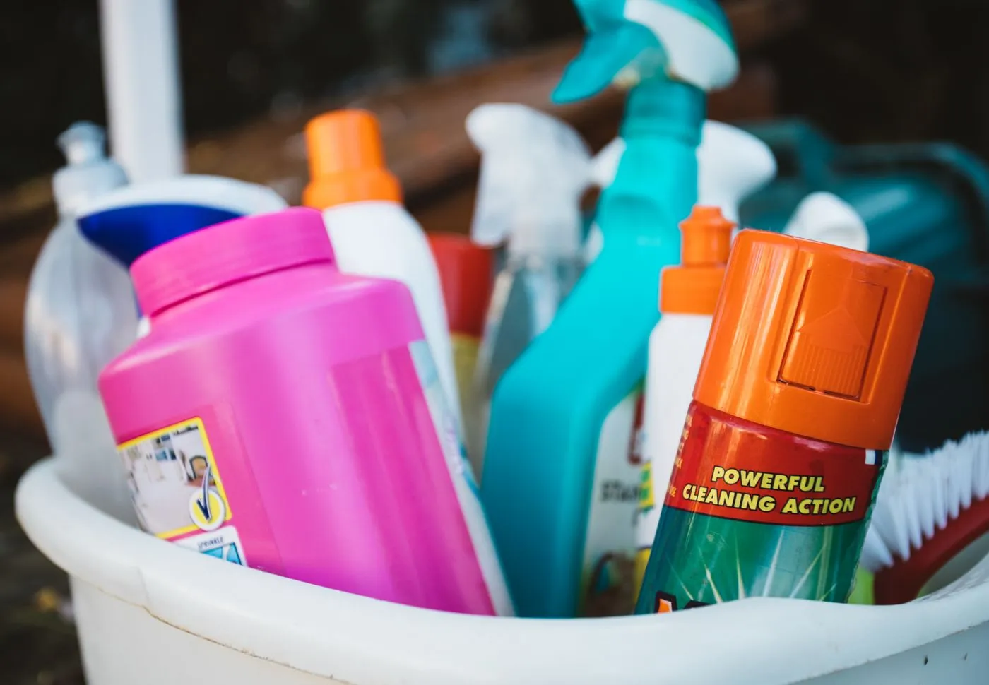 Close-up of cleaning sprays, a bleach bottle and a scrubbing brush standing in a white plastic bucket