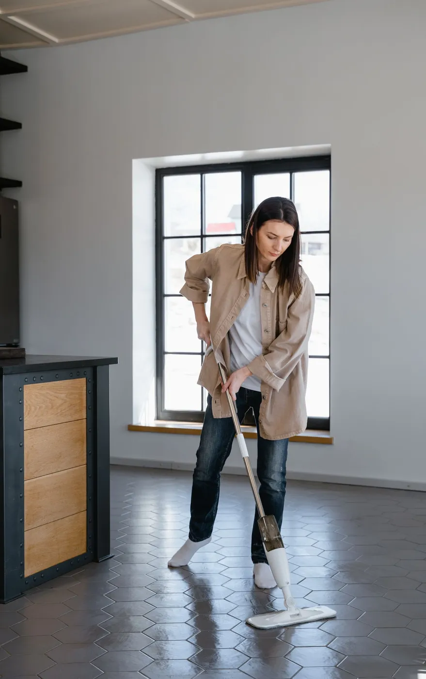 Woman in a beige jacket mopping a dark hexagonal tiled floor in a bare room with a tall window behind her