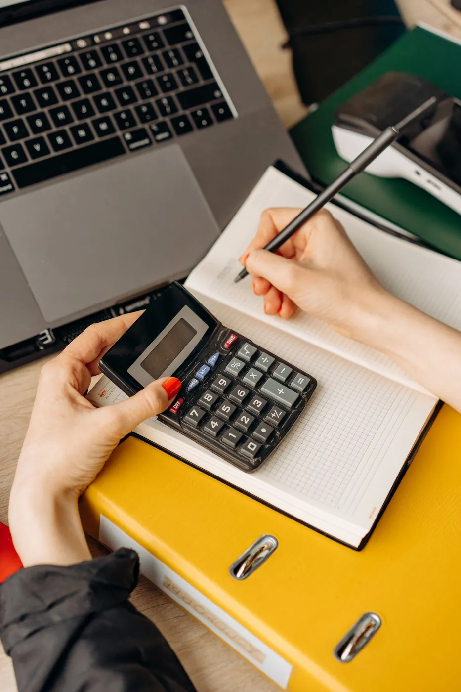 Hands holding a small black calculator over an open grid notebook beside a laptop and a yellow lever arch file