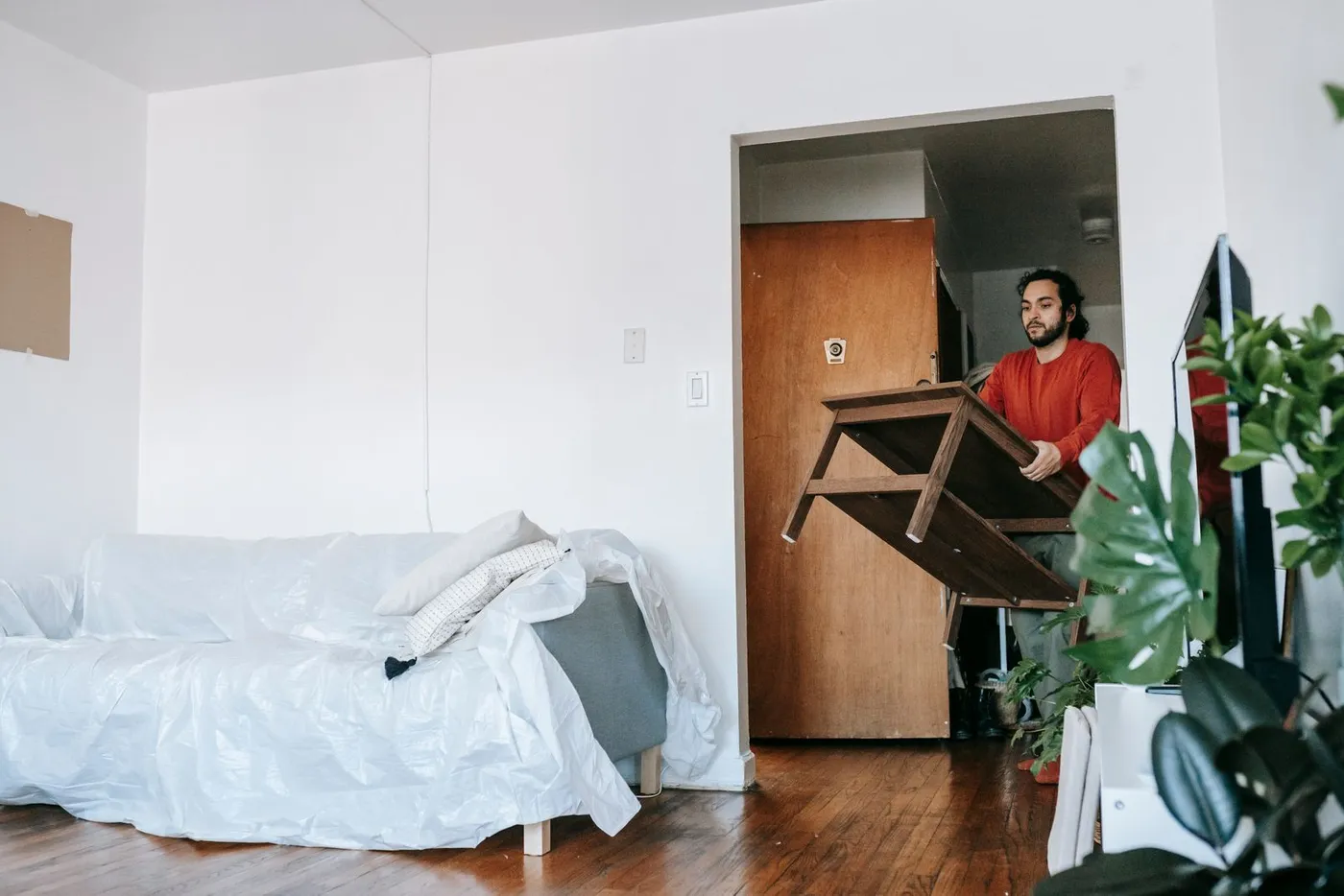 Man in a red top carrying a dark wooden table through a doorway past a sofa wrapped in protective plastic
