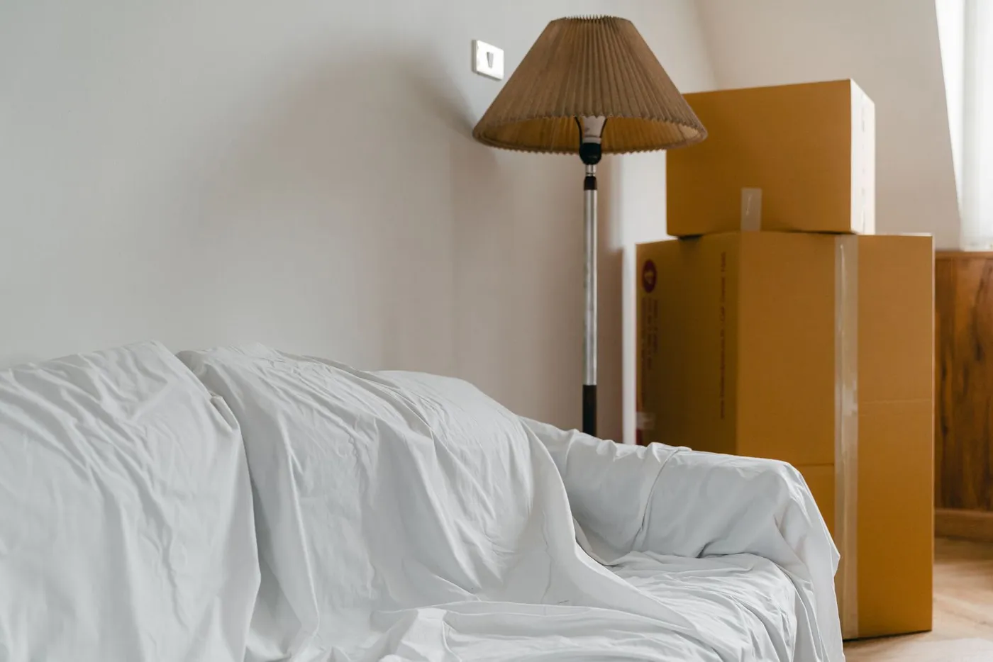 Sofa covered with a white dust sheet beside a floor lamp and two stacked cardboard moving cartons in a bare room