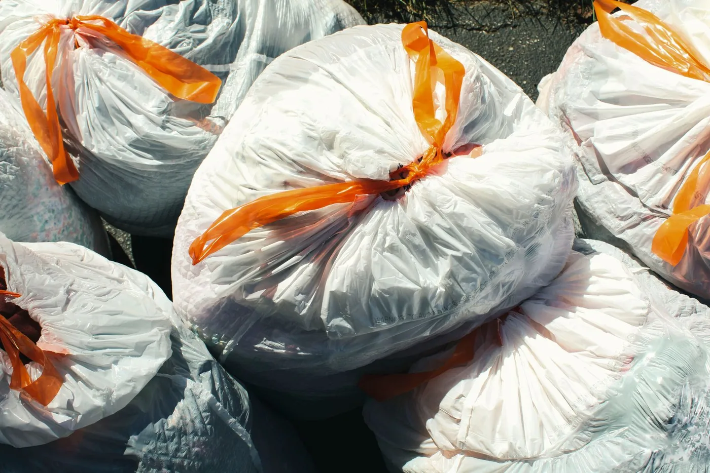Pile of filled white refuse sacks tied with bright orange ribbon ties, stacked on tarmac in strong sunlight