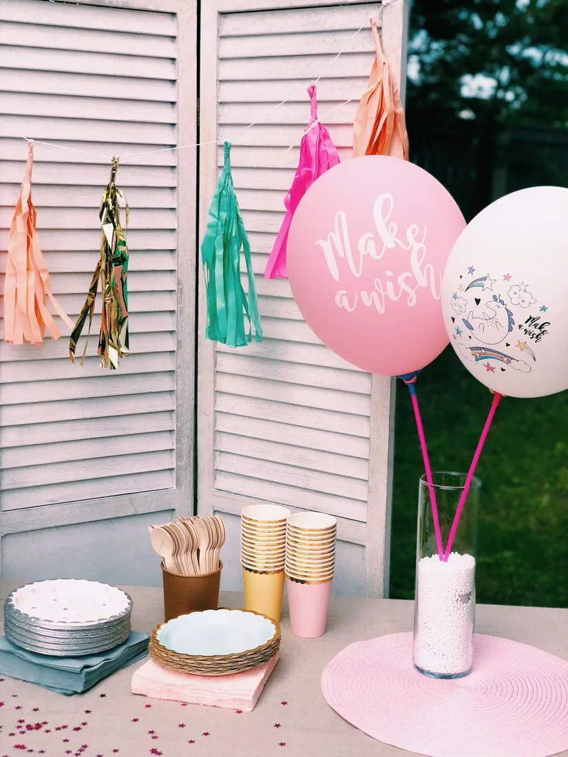 Outdoor party table with stacked gold and pink paper cups, layered paper plates, wooden cutlery in a tub, folded napkins, hanging tassel garlands and two balloons