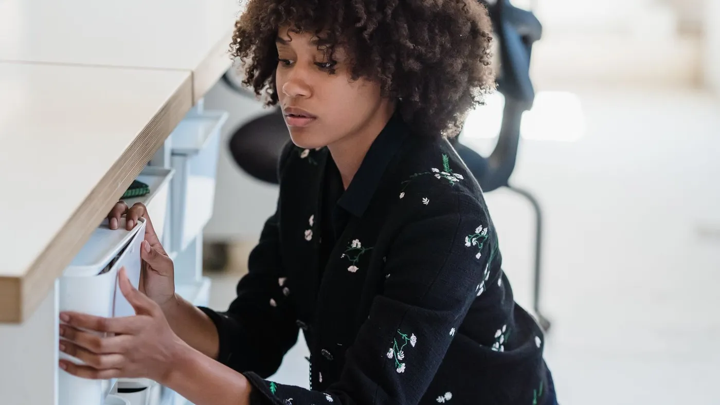 Person in a dark floral jacket crouching beside a white under-desk pedestal drawer unit, pulling the drawer open with both hands