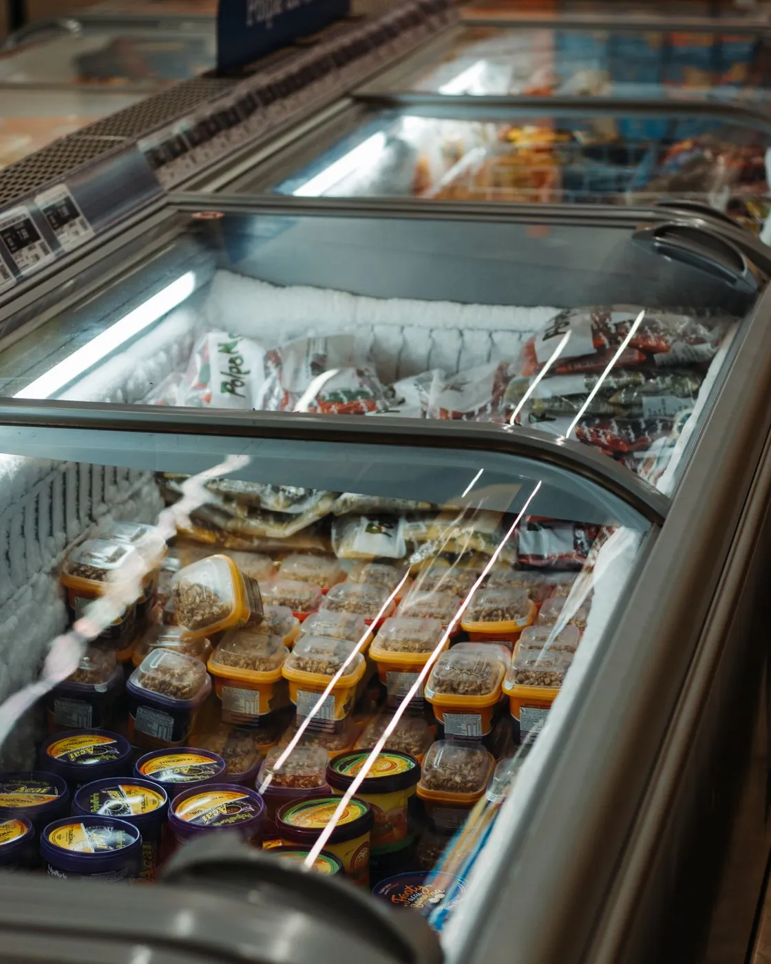 Row of glass-lid commercial chest display freezers stocked with frozen products, with thick white frost built up along the interior walls
