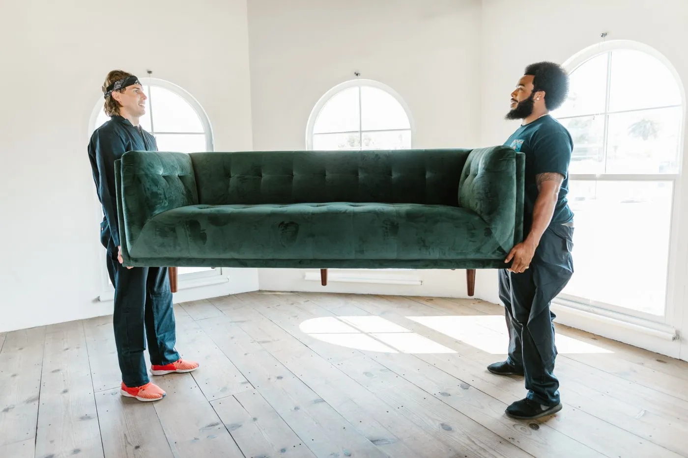 Two workers in dark overalls carrying a green velvet sofa across an empty room with arched windows