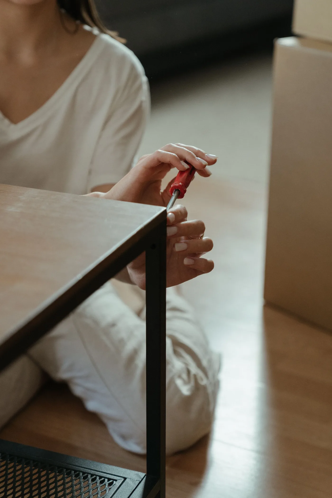 Person sitting on the floor using a red screwdriver on the metal frame of a wooden table, with a moving box beside them