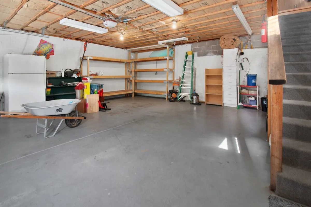 Cleared garage with a swept concrete floor, empty timber shelving along the wall, a folded ladder, a wheelbarrow and a single upright freezer