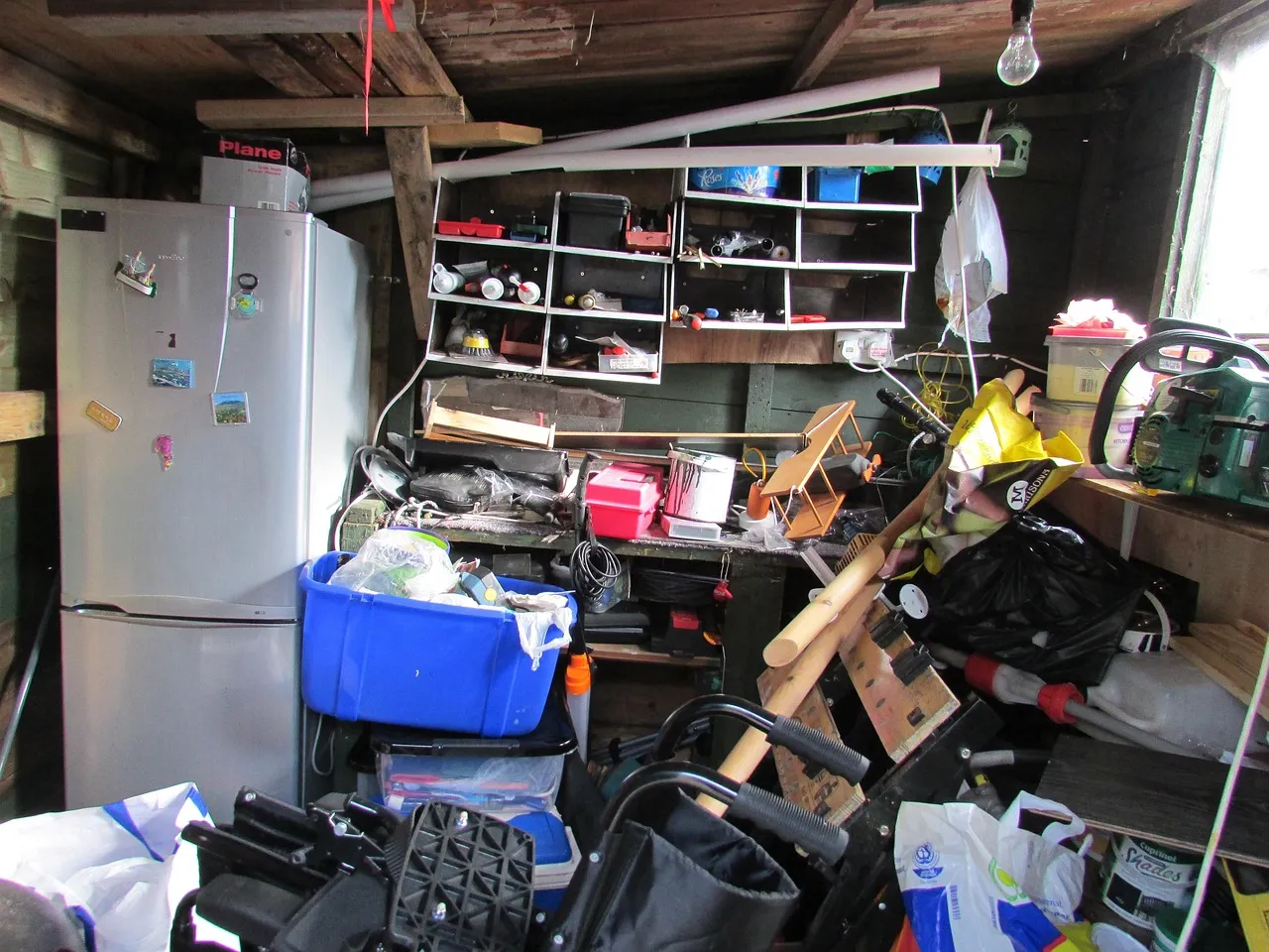 Garage store crammed floor to ceiling with an old fridge, a cluttered workbench, stacked plastic crates, bags and loose timber