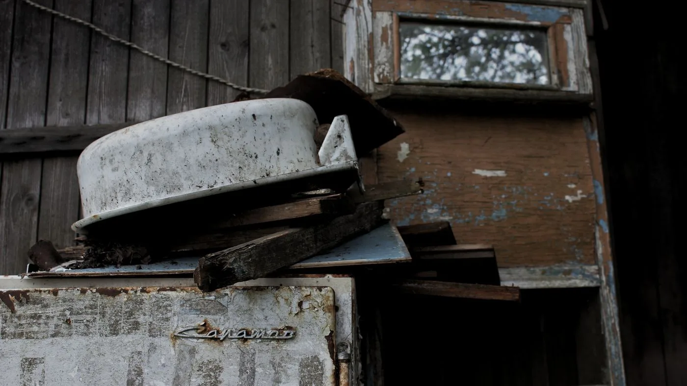 Discarded white enamel appliance casing tipped on its side among broken timber against a weathered wooden wall