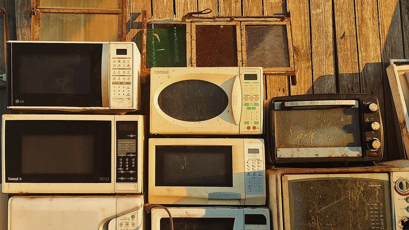 Stack of old microwave ovens and countertop cookers with dusty glass doors piled against a wooden plank wall
