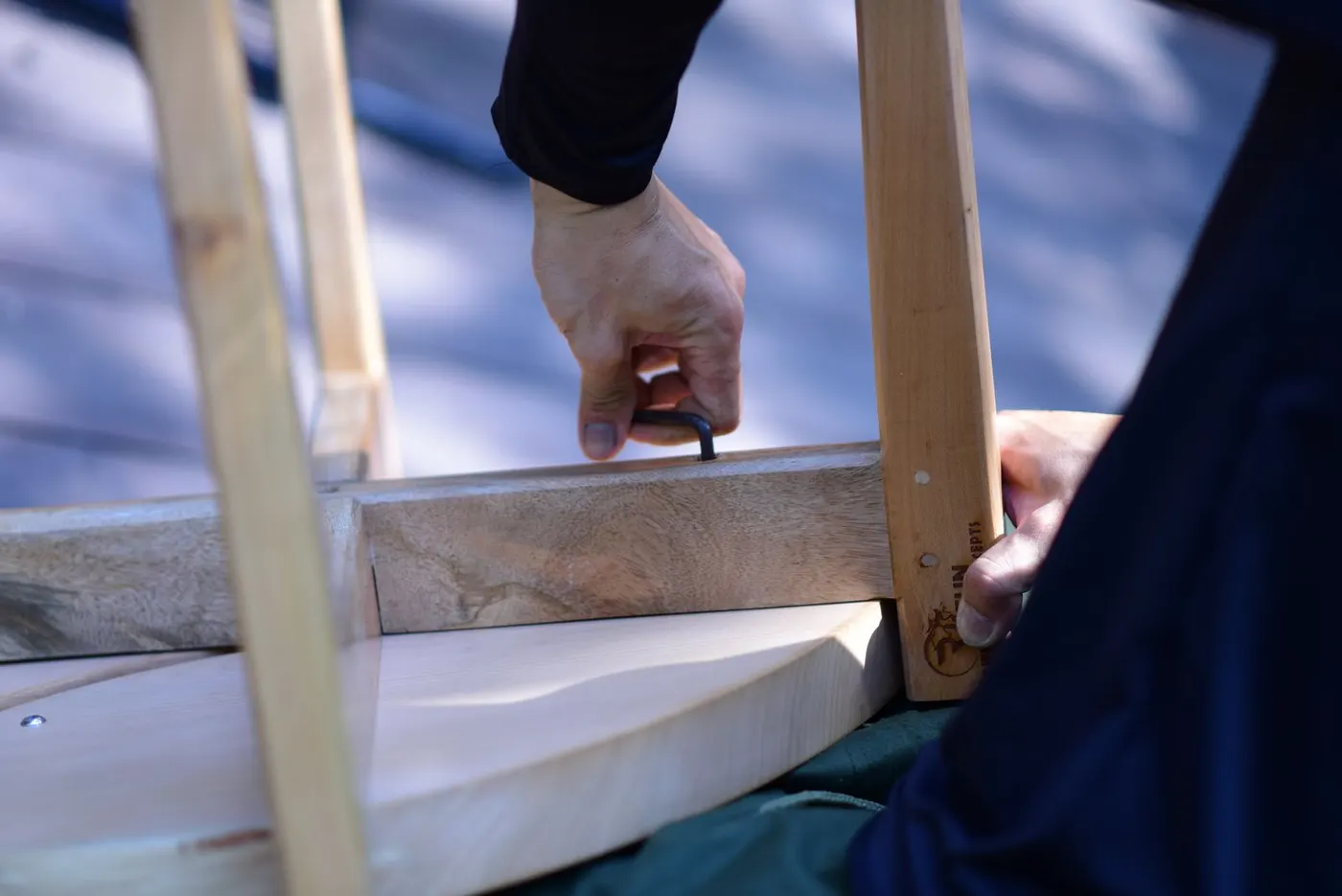 Close-up of a hand using a black hex key on the underside of a light wooden table frame while another hand steadies the leg