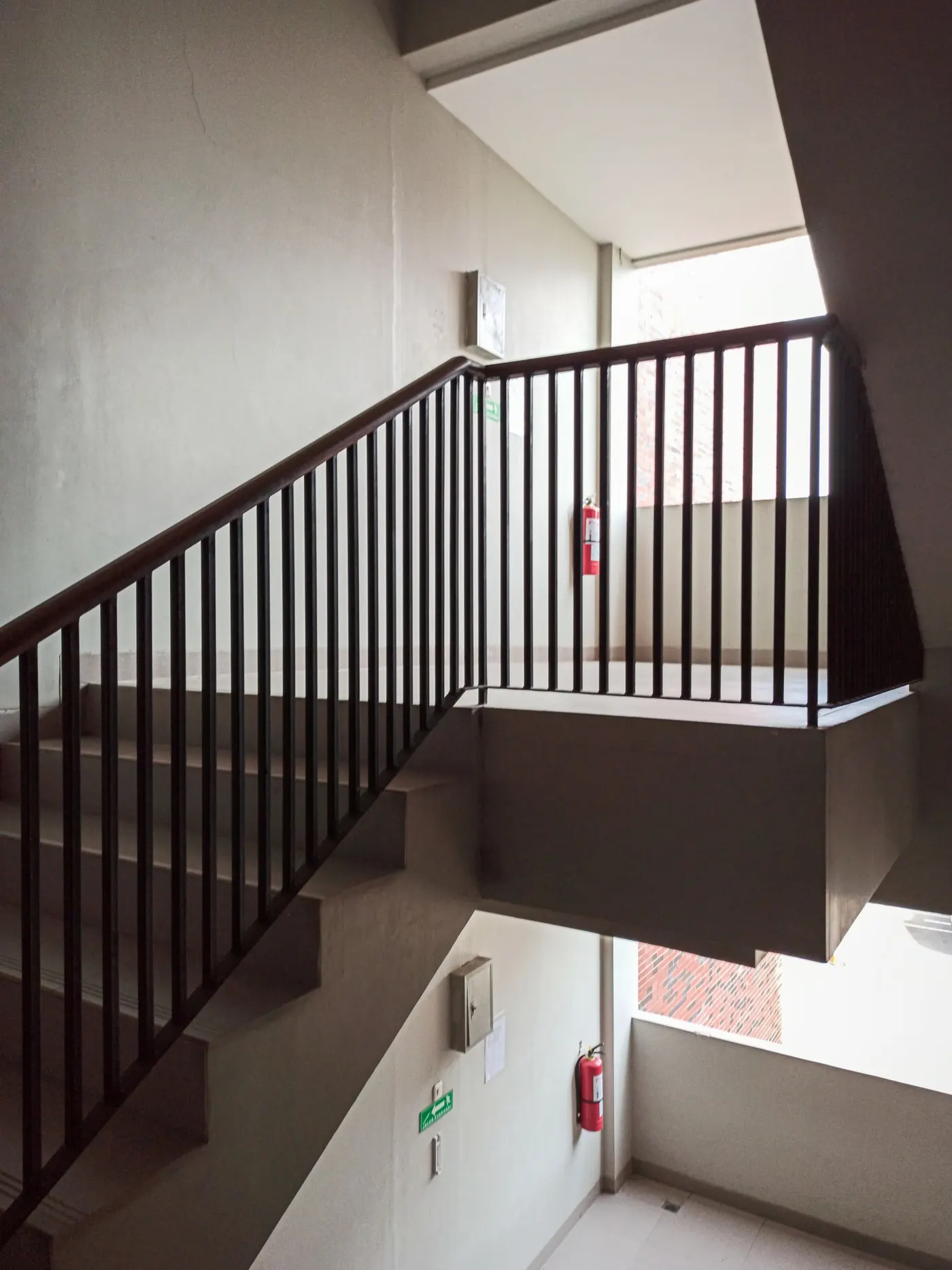 Concrete stairwell in a residential building with a dark wooden handrail, red fire extinguishers mounted on the landing walls and a green exit sign