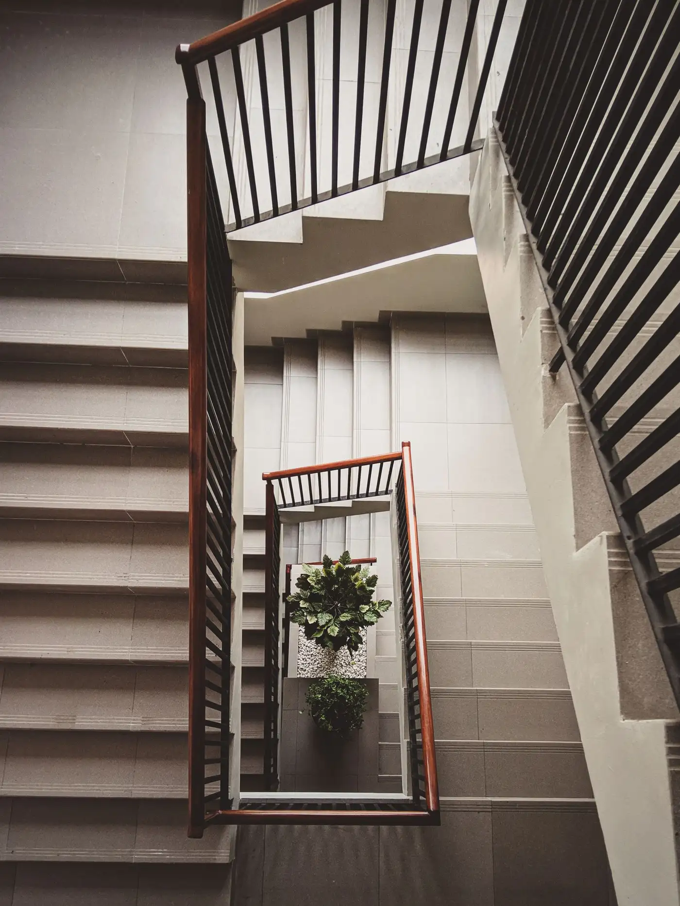 View looking down a residential stairwell with wooden handrails, concrete steps and potted plants at the base, showing the tight rectangular turn between flights