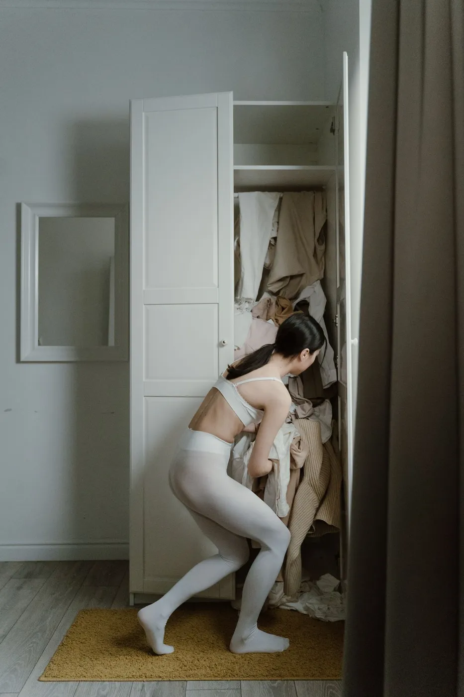 Person in white leggings pushing against an overstuffed wardrobe with clothing spilling out of the doors