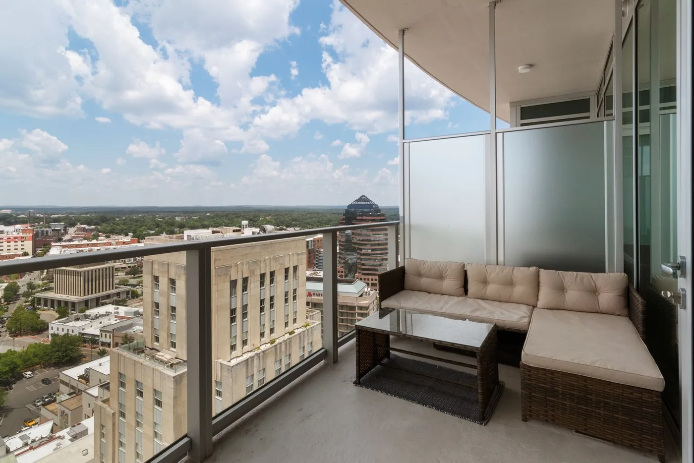 High-floor apartment balcony with a rattan corner sofa, beige cushions and a glass-topped table, overlooking office buildings and rooftops under a cloudy sky