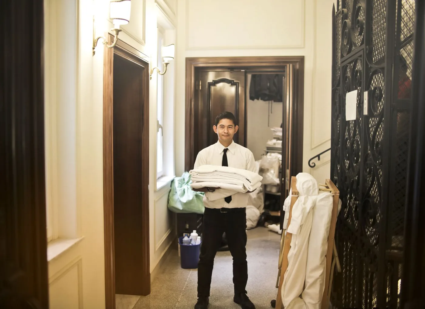 Housekeeping staff member in a white shirt and black tie carrying a stack of folded white linen along a corridor, with a laundry trolley and cleaning supplies behind