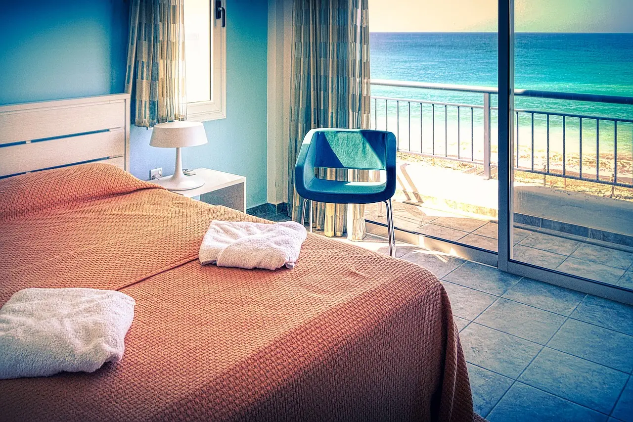 Hotel bedroom with an orange bedspread, folded white towels and a chair beside a balcony overlooking the sea