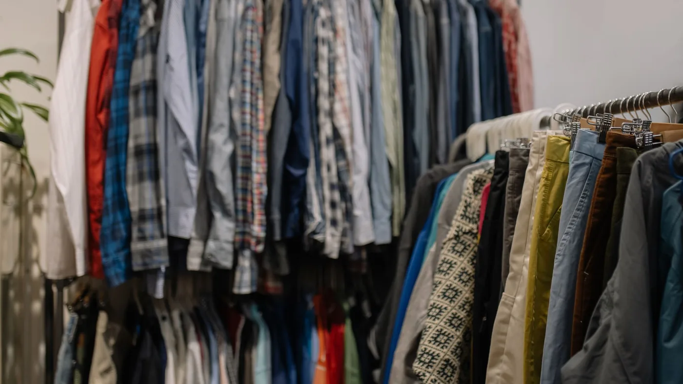 Two packed rails of second-hand shirts, jackets and trousers on hangers against a plain wall