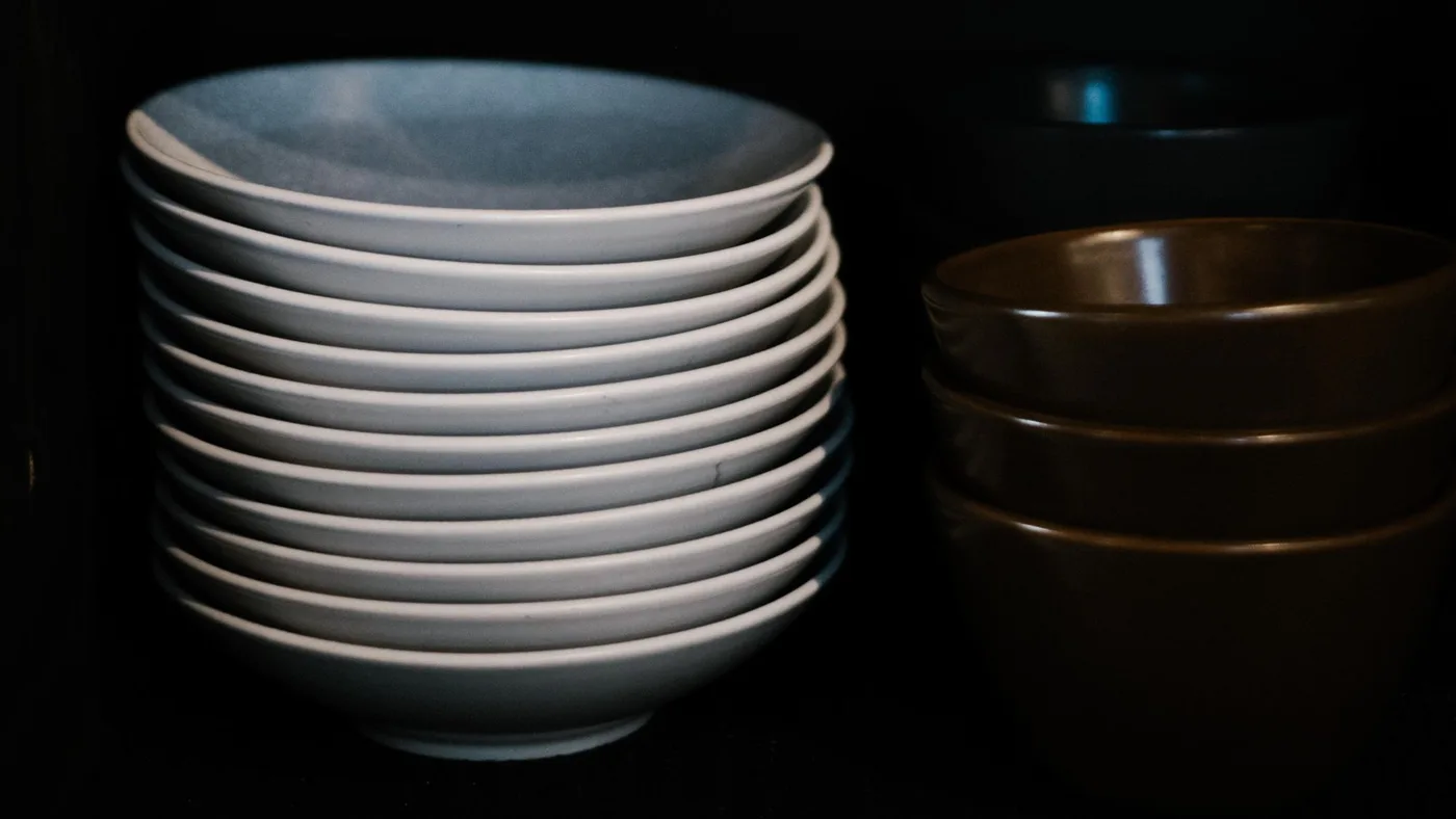 Tall stack of white bowls beside three brown bowls on a shelf inside a dark kitchen cupboard