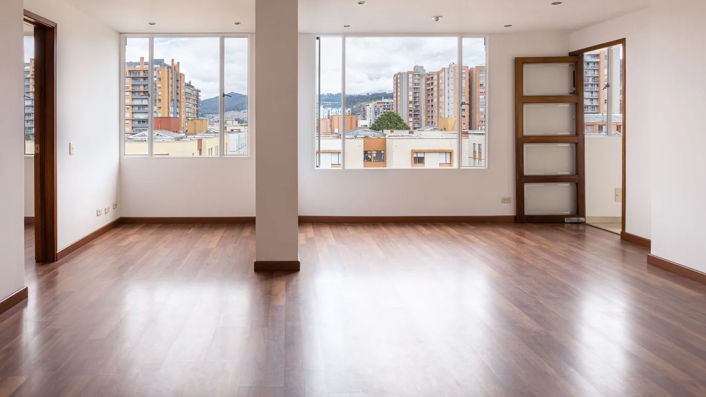 Emptied apartment living room with polished wooden flooring, a central pillar and two large windows looking out over nearby apartment blocks