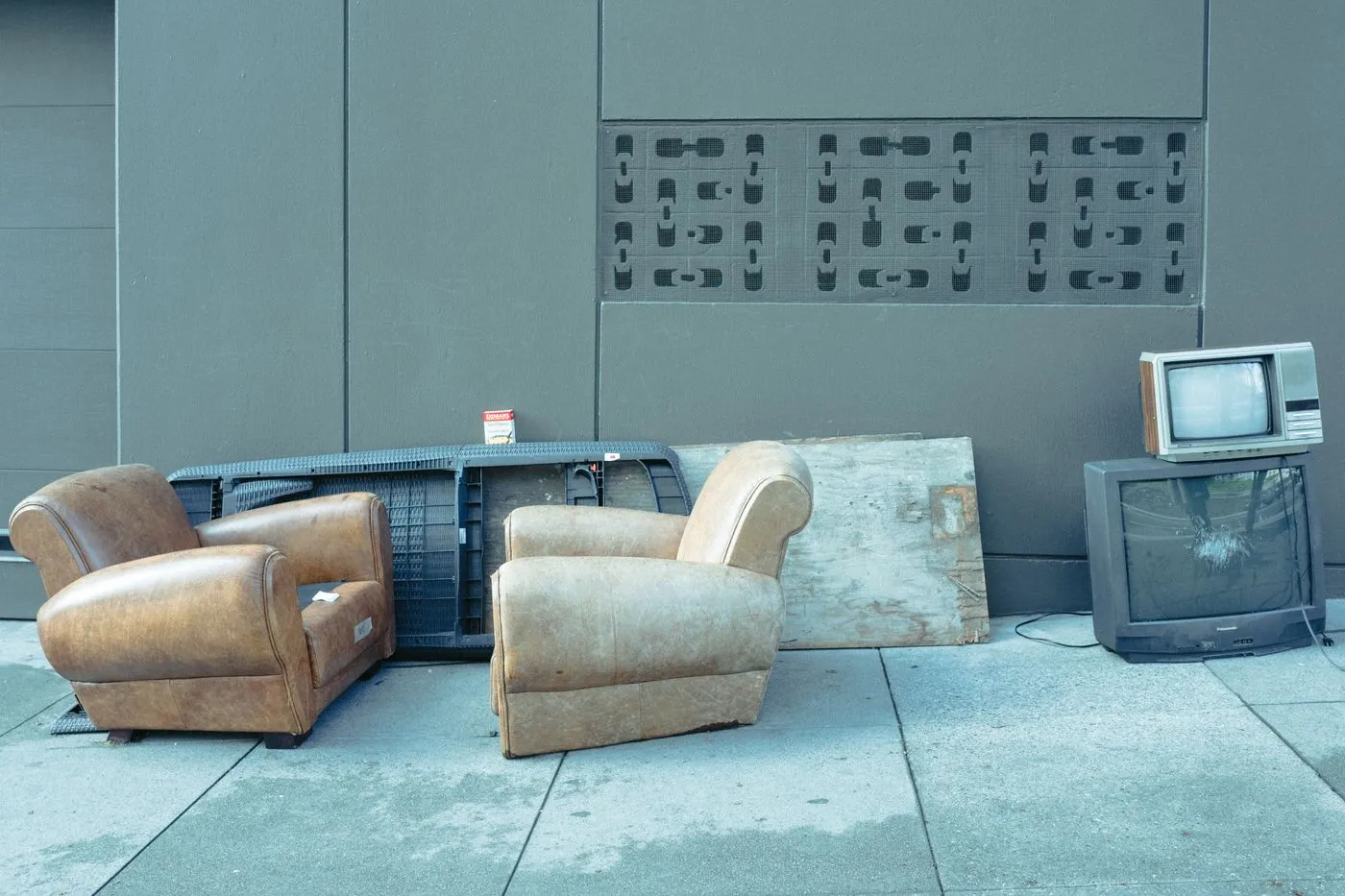 Two worn leather armchairs and two old television sets left on a pavement against a grey wall