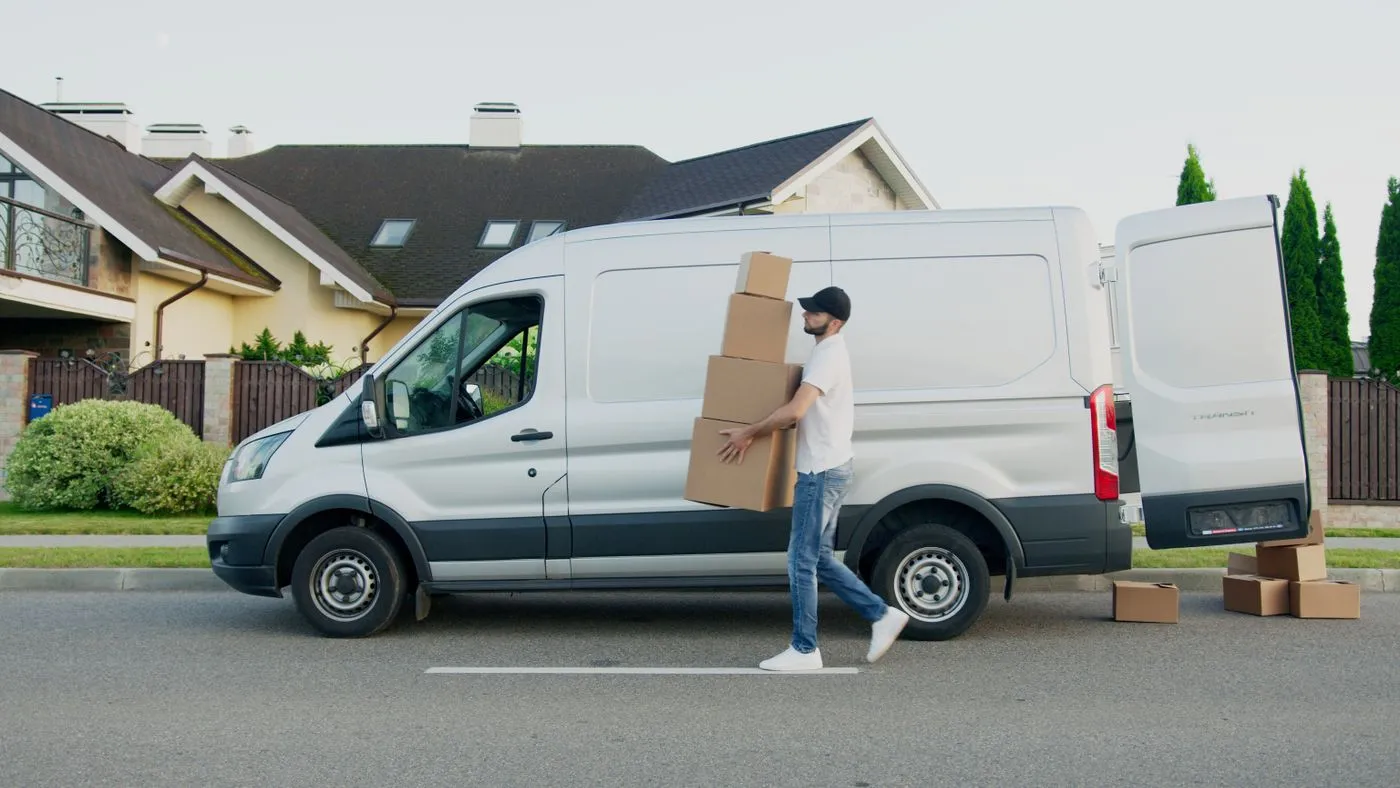 Man carrying a stack of cardboard boxes towards the open rear doors of a white van parked outside a house