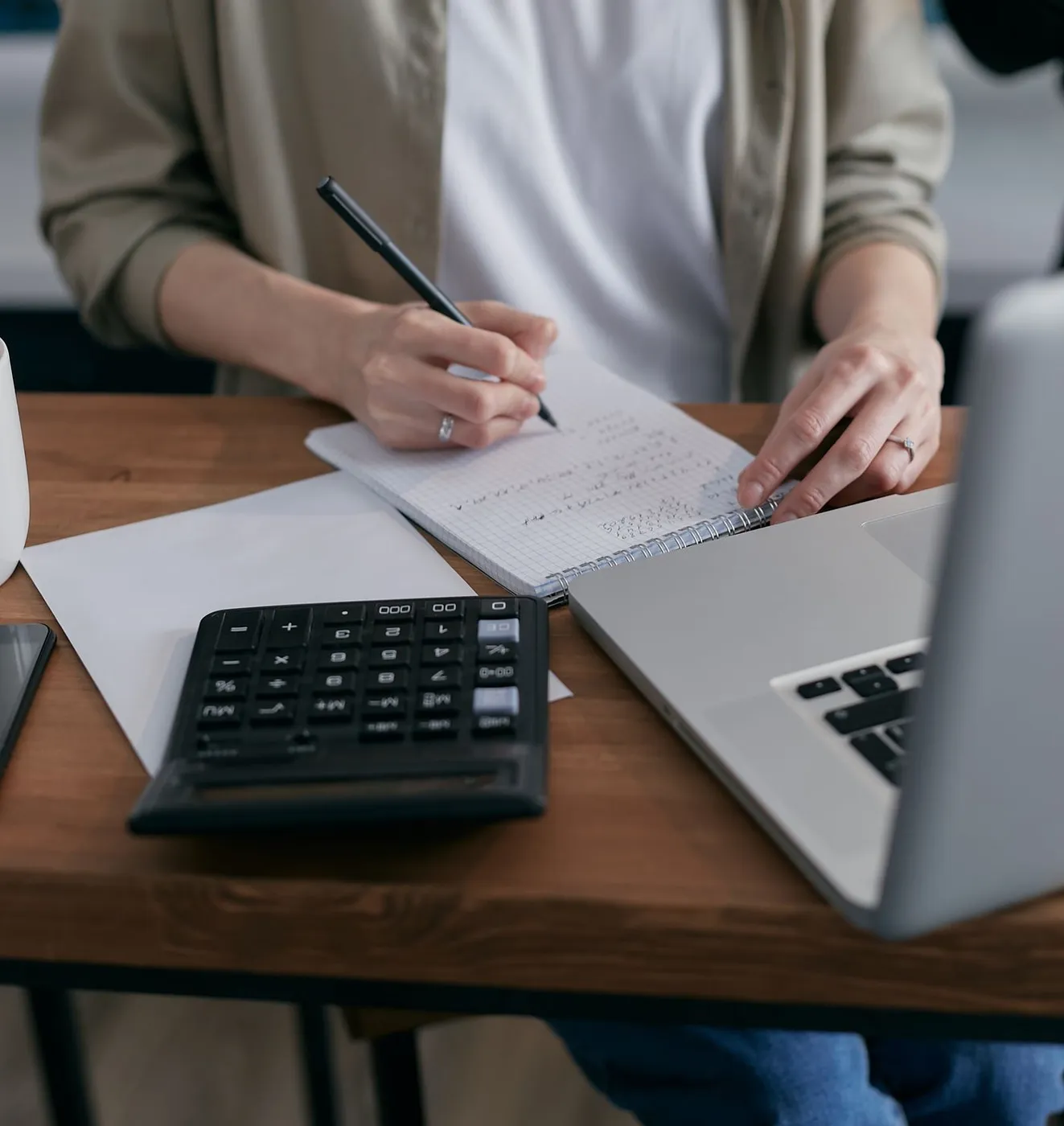 Person writing figures in a spiral notebook beside a black desk calculator and an open laptop