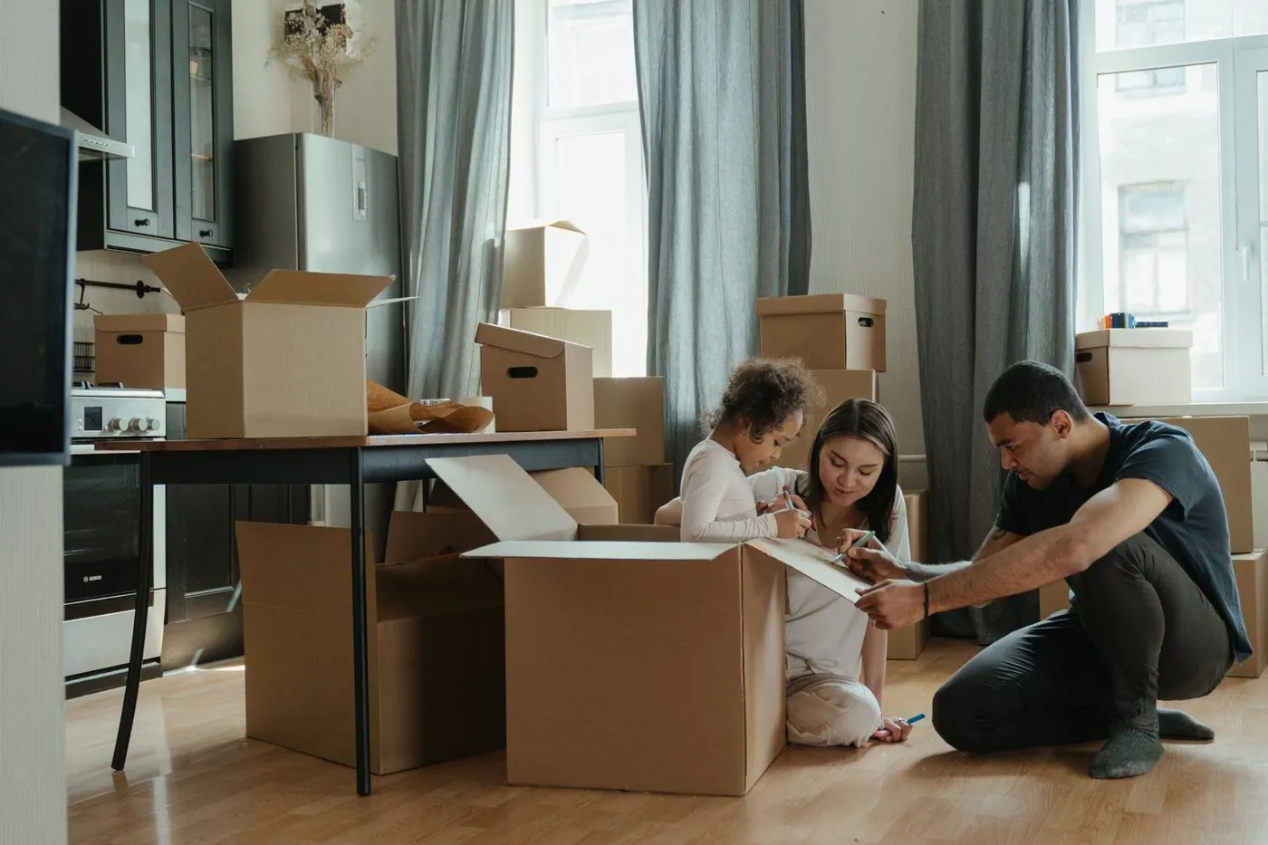 Family sorting through open cardboard boxes on the floor of a kitchen and dining area during a move