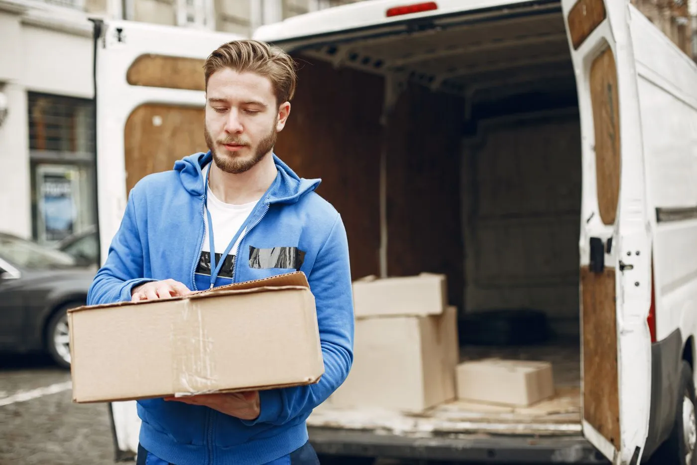 Man in a blue hooded top carrying a cardboard box towards the open rear doors of a white van loaded with boxes