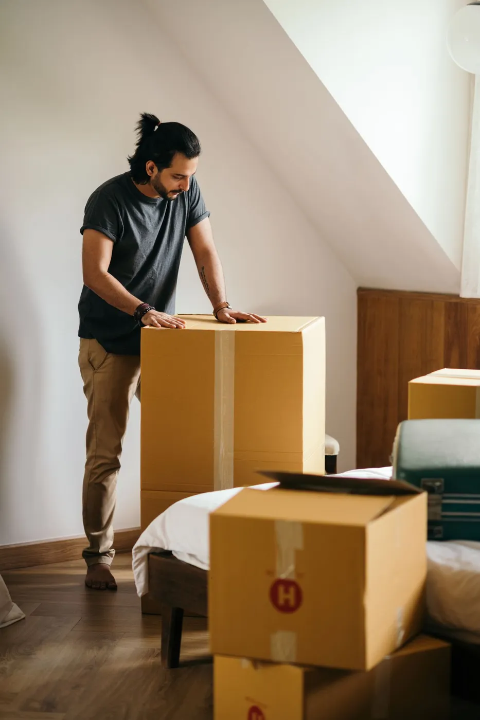 Man taping a large cardboard box closed in a bedroom with a suitcase and more packed boxes beside the bed