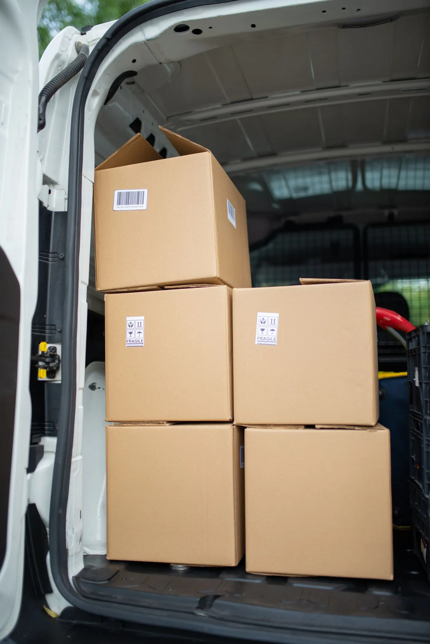 Stacked sealed cardboard boxes with fragile labels loaded into the open rear of a white van, with a plastic crate visible behind them