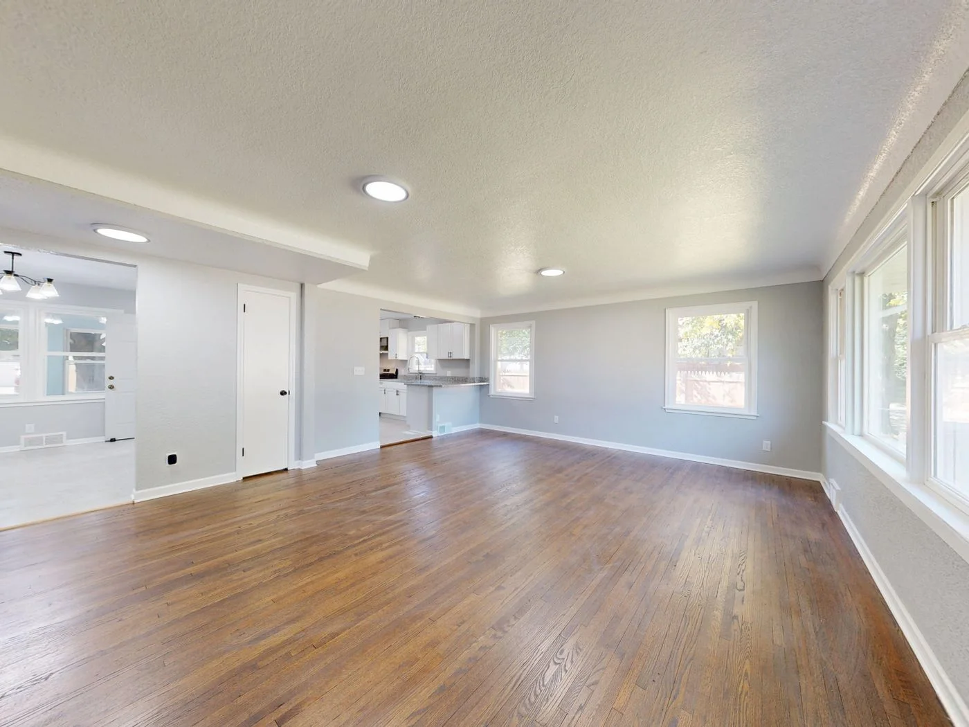 Emptied living room with dark hardwood flooring, pale grey walls, recessed ceiling lights and daylight coming through a bank of windows