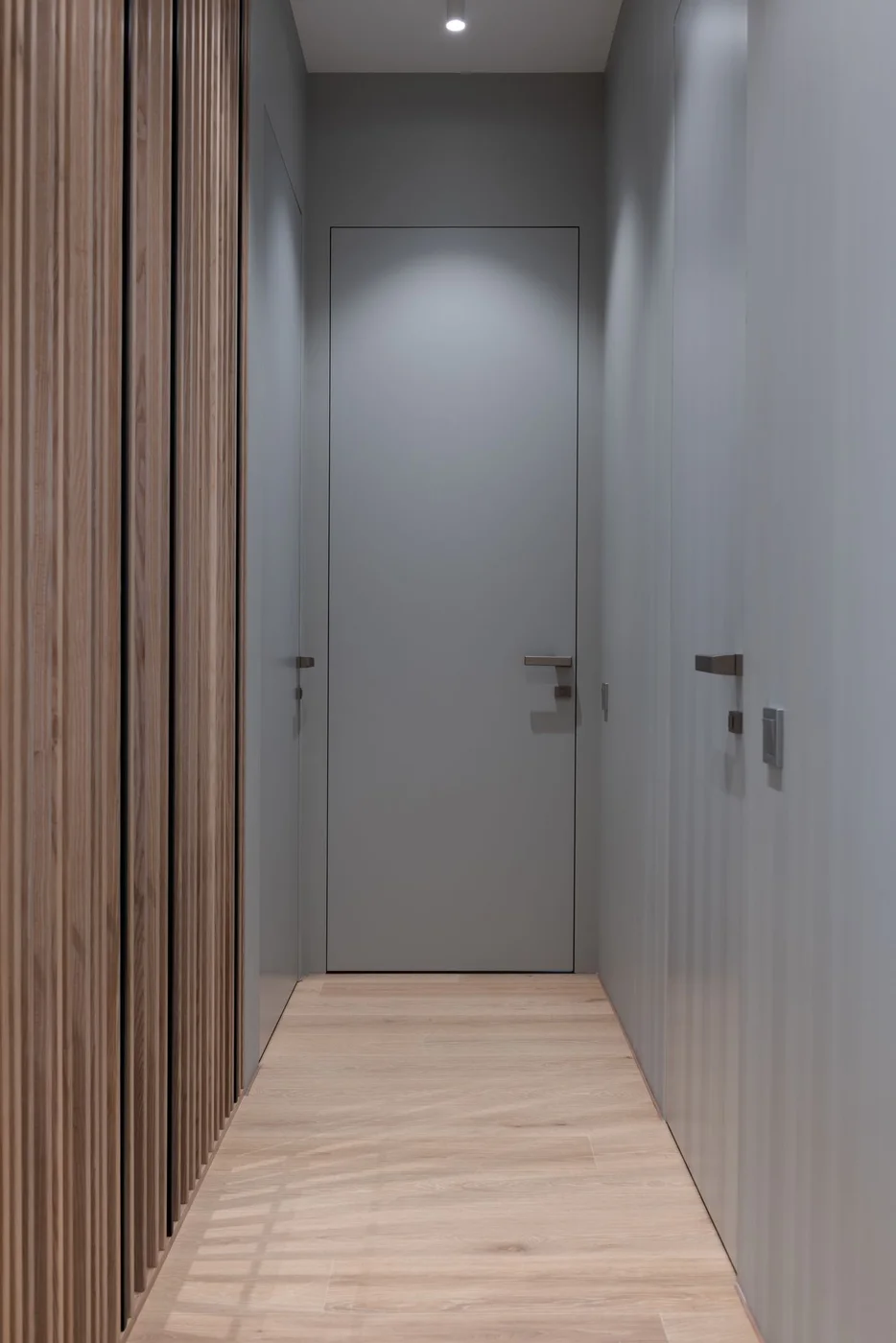 Narrow apartment hallway with a slatted wood wall panel on one side, a flush grey door at the end and pale timber flooring