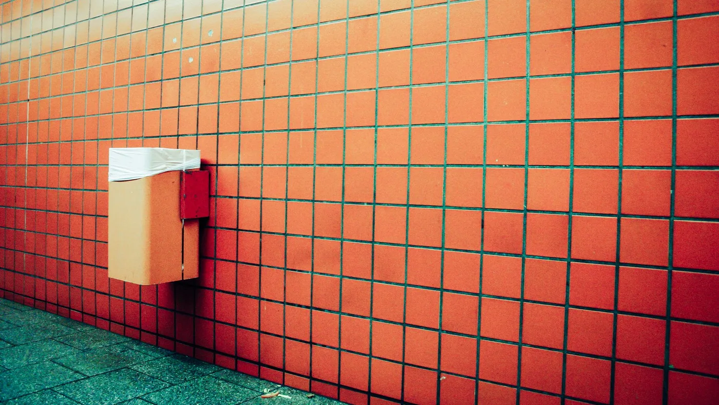 Small wall-mounted waste bin fixed to an orange tiled wall in a covered walkway, wrapped with a loose plastic liner