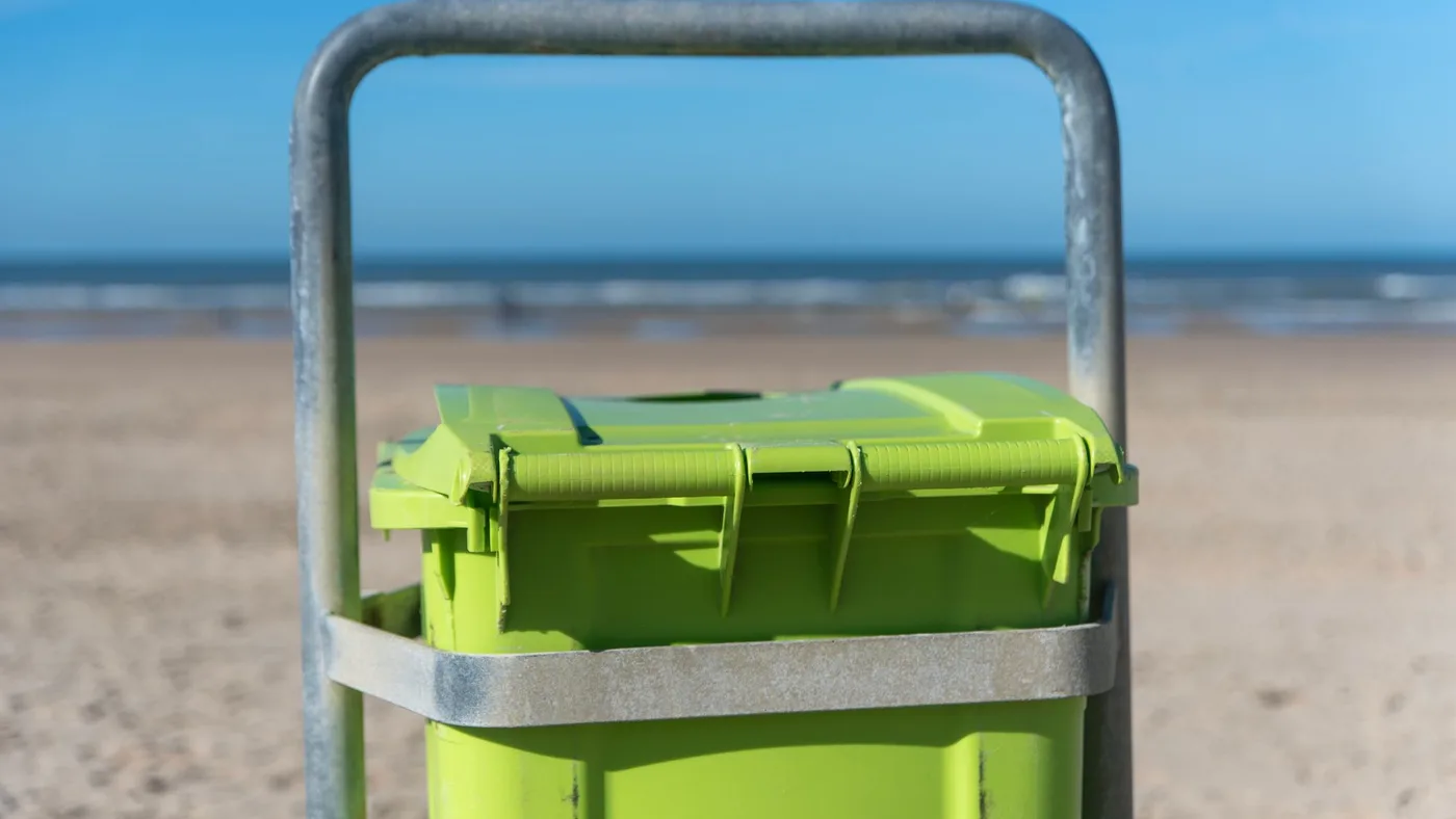 Bright green outdoor bin fixed to a metal frame, seen close up with a sandy shoreline and the sea blurred behind it