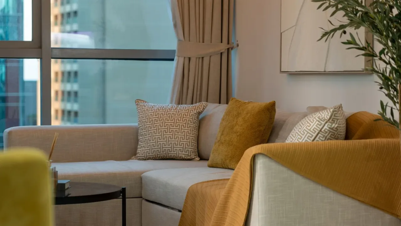 Corner sofa with neatly arranged patterned and mustard cushions and a folded throw beside a tall window in a tower apartment