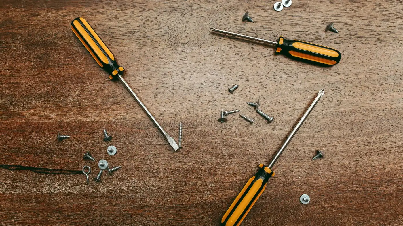 Two yellow-handled screwdrivers lying on a dark wooden floor beside scattered wood screws, washers and a hex key